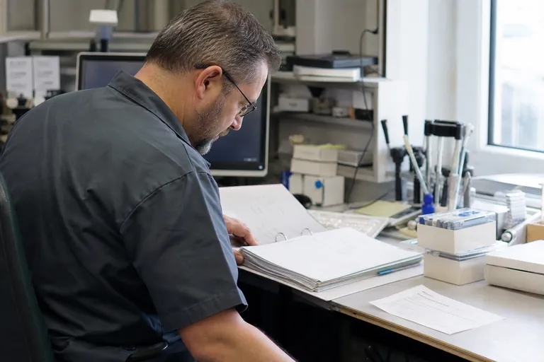 Zahntechniker vergleicht am Arbeitsplatz Auftragsunterlagen, Materialschachteln und einen Lieferschein.